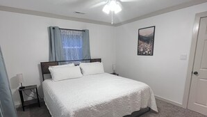 Ein Schlafzimmer mit einem Einzelbett, einem Deckenventilator, einem Nachttisch, einer Lampe, einem Fenster mit Vorhängen und einer Tür.