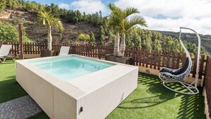 A rectangular hot tub with a textured exterior, surrounded by a wooden fence, a hanging chair, and a view of a hilly landscape. - Vacation Home 'Añaterve' with Sea View, Private Pool and Wi-Fi (Arico)