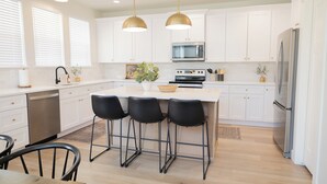 Une cuisine moderne avec des meubles blancs, un îlot central doté de tabourets de bar, des appareils en acier inoxydable et un éclairage suspendu.