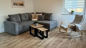Una sala de estar con un sofá gris, dos mesitas de madera, un sillón mecedora y una ventana con persianas.