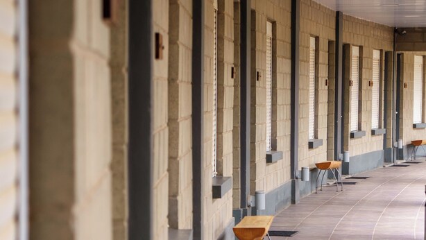 Um longo corredor com várias janelas e portas em um dos lados, e algumas mesas e cadeiras no centro.