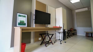 Um espaço de escritório moderno com uma mesa grande, uma televisão de tela plana montada, um certificado emoldurado e uma lixeira vermelha.