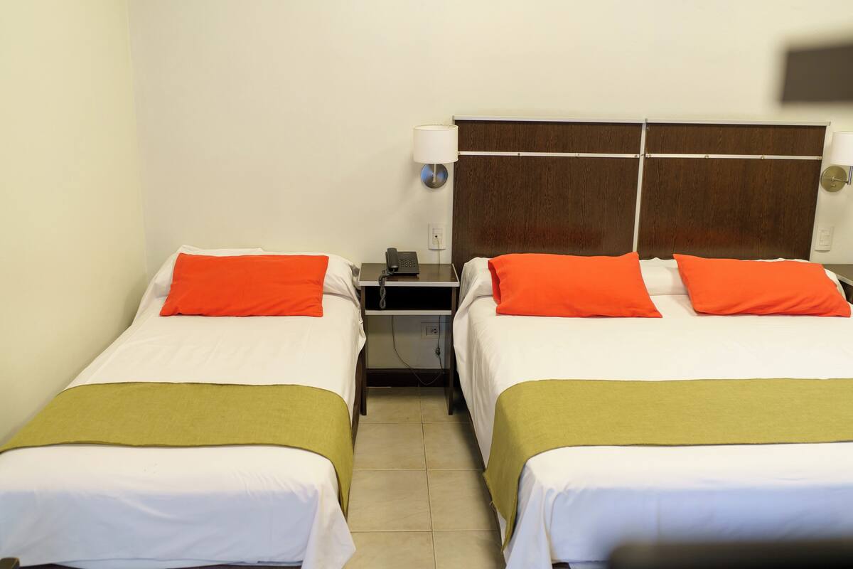 A hotel room with two single beds, each with an orange pillow and a green bedspread. There is a nightstand with a phone and a lamp between the beds.