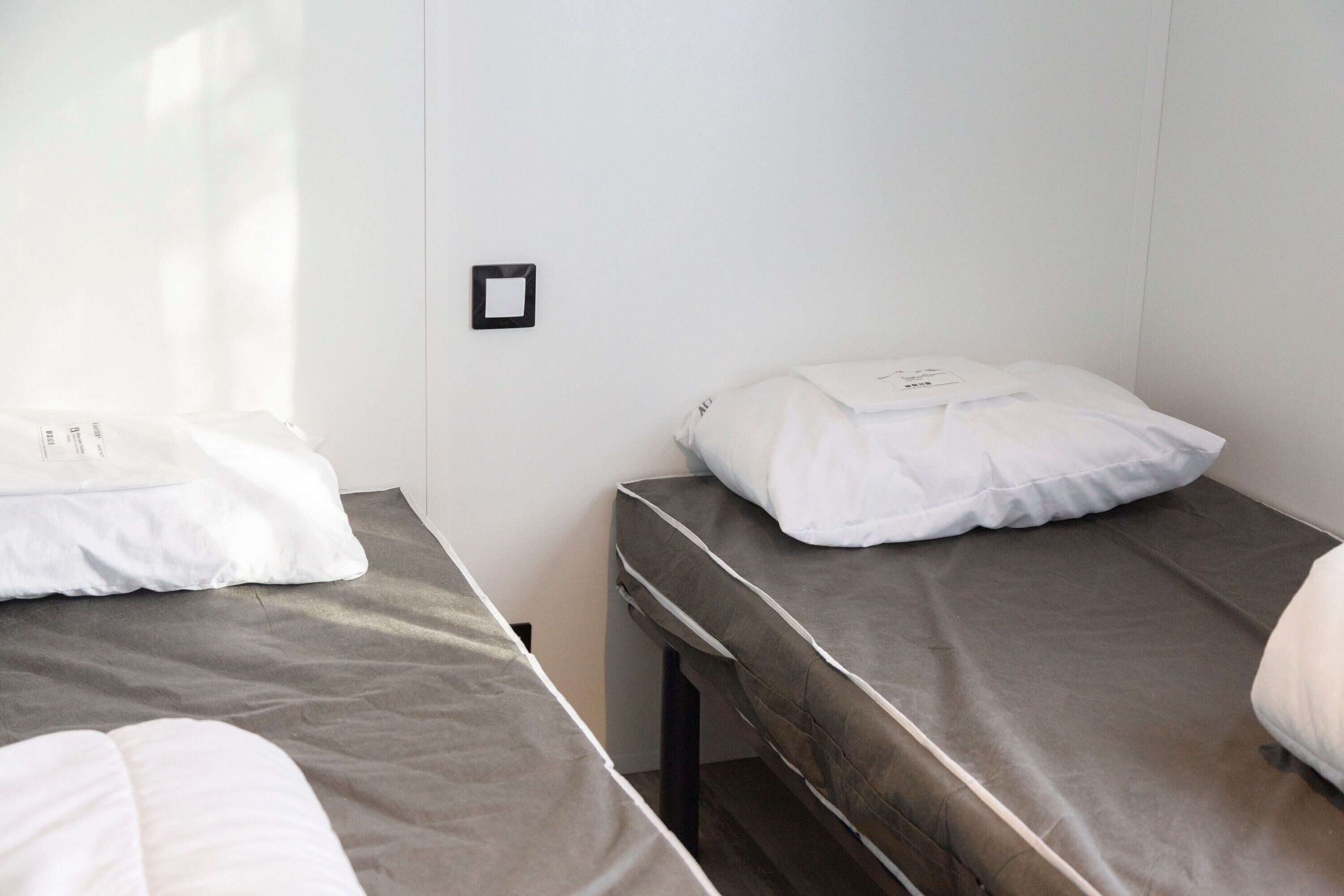 Deux lits simples avec du linge de lit blanc, une petite table et un luminaire fixé au mur, dans une pièce aux murs blancs.