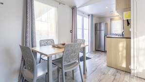 A modern kitchen with a refrigerator, wooden cabinets, and a dining area with a table and chairs. - Mobile Home 'Sur Pont-Aven' with Private Terrace, Garden, and Wi-Fi (Pont-Aven)