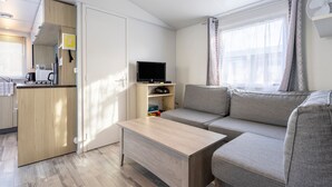 A compact living space with a kitchenette, a sofa, a flat-screen TV, and a window with curtains. - Mobile Home 'Sur Pont-Aven' with Private Terrace, Garden, and Wi-Fi (Pont-Aven)