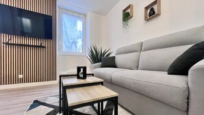 Una sala de estar moderna con un sofá gris, una pequeña mesa de centro de madera, un televisor montado en una pared de listones y una ventana con vistas a un cielo azul.