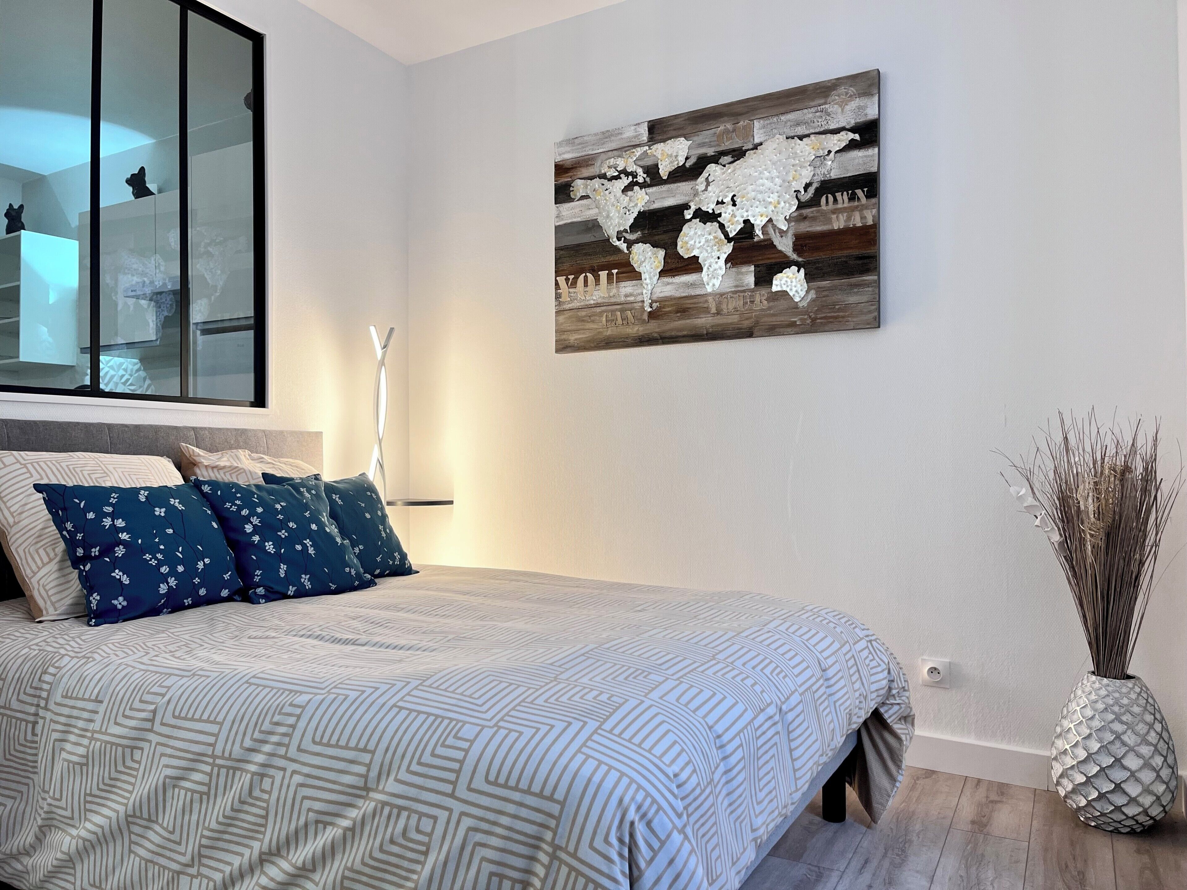 Un dormitorio con una cama, un cuadro de un mapa del mundo, una lámpara de pie alta y un jarrón decorativo.