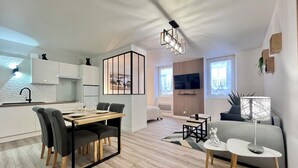 Un apartamento moderno con cocina, comedor y sala de estar. Tiene piso de madera, una mesa de comedor con sillas, un sofá y un televisor montado en la pared.