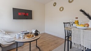 A cozy living room with a flat-screen TV displaying "NETFLIX," a round coffee table with books and pastries, a dining area with a table set for two, and decorative wall art. - Lovely flat near Gatwick Airport with free parking  (Surrey)