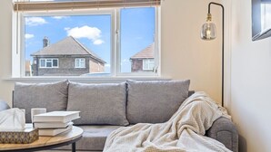 A cozy living room with a grey sofa, a round coffee table with books and a tissue box, a blanket draped over the sofa, and a window with a view of houses. - Lovely flat near Gatwick Airport with free parking  (Surrey)