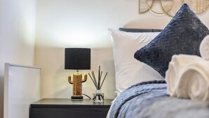 A neatly made bed with a blue pillow and a white pillow, a gold lamp, and a small glass container with black sticks on a dark wooden nightstand. - Lovely flat near Gatwick Airport with free parking  (Surrey)