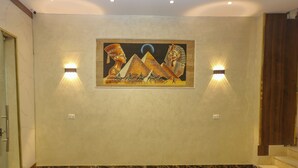 Um corredor com piso de mármore, uma pintura embutida na parede retratando figuras egípcias, e duas luminárias fixadas na parede.