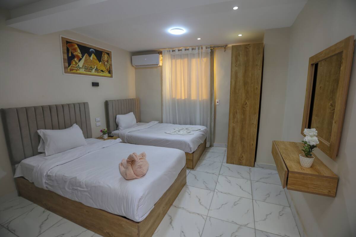 A hotel room with two beds, a wooden headboard, a small table with a flower, and a painting on the wall.