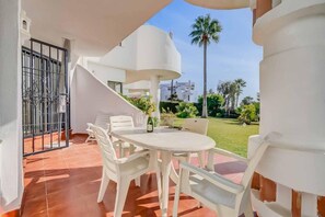 A terrace with a white table and chairs, a bottle of champagne, and a view of a white building and palm trees.