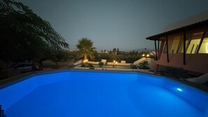 A swimming pool with a wooden deck in the foreground, surrounded by a landscaped garden with palm trees and a building with large windows in the background.