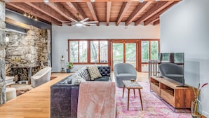 A spacious living room with a stone fireplace, wooden ceiling beams, large windows, and a modern entertainment unit. - Stone Cabin on Valley View: Fire Pit + Hot Tub (Harpers Ferry)