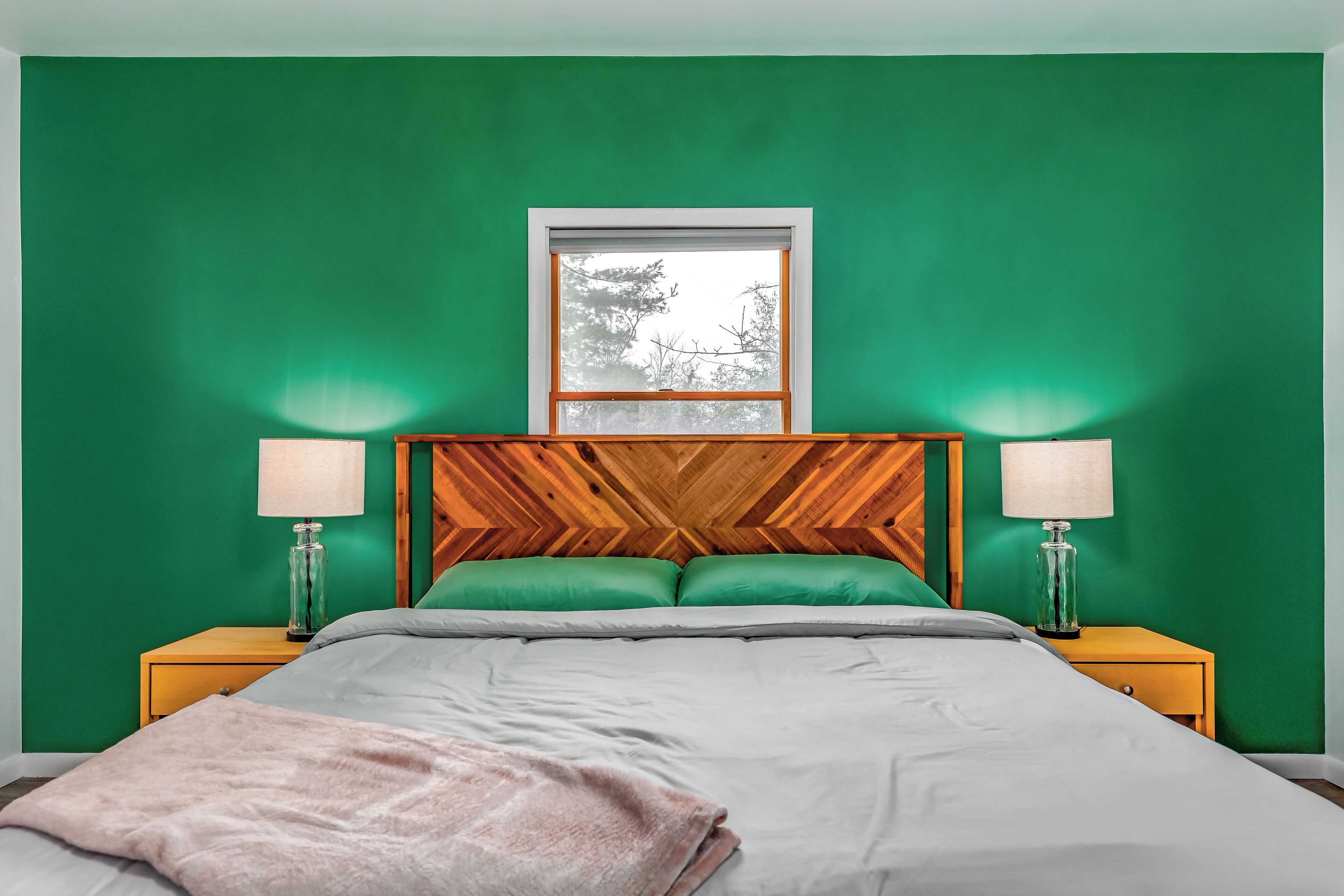 Une chambre avec un mur vert, une tête de lit en bois, deux tables de chevet avec des lampes, un lit avec une couette grise et blanche, et une fenêtre donnant sur des arbres.