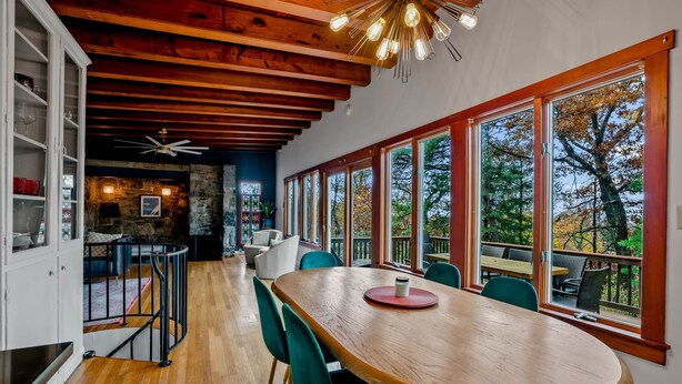 A spacious dining area with a wooden table and chairs, large windows with a view of trees, and a stone wall in the background. - Stone Cabin on Valley View: Fire Pit + Hot Tub (Harpers Ferry)