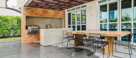 An outdoor patio with a brick barbecue, a wooden table with chairs, and large windows.