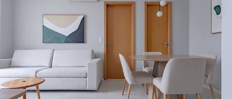 Een moderne woonkamer met een bank, een ronde eettafel en stoelen. Er is een houten deur en een lijst met een abstract schilderij aan de muur.