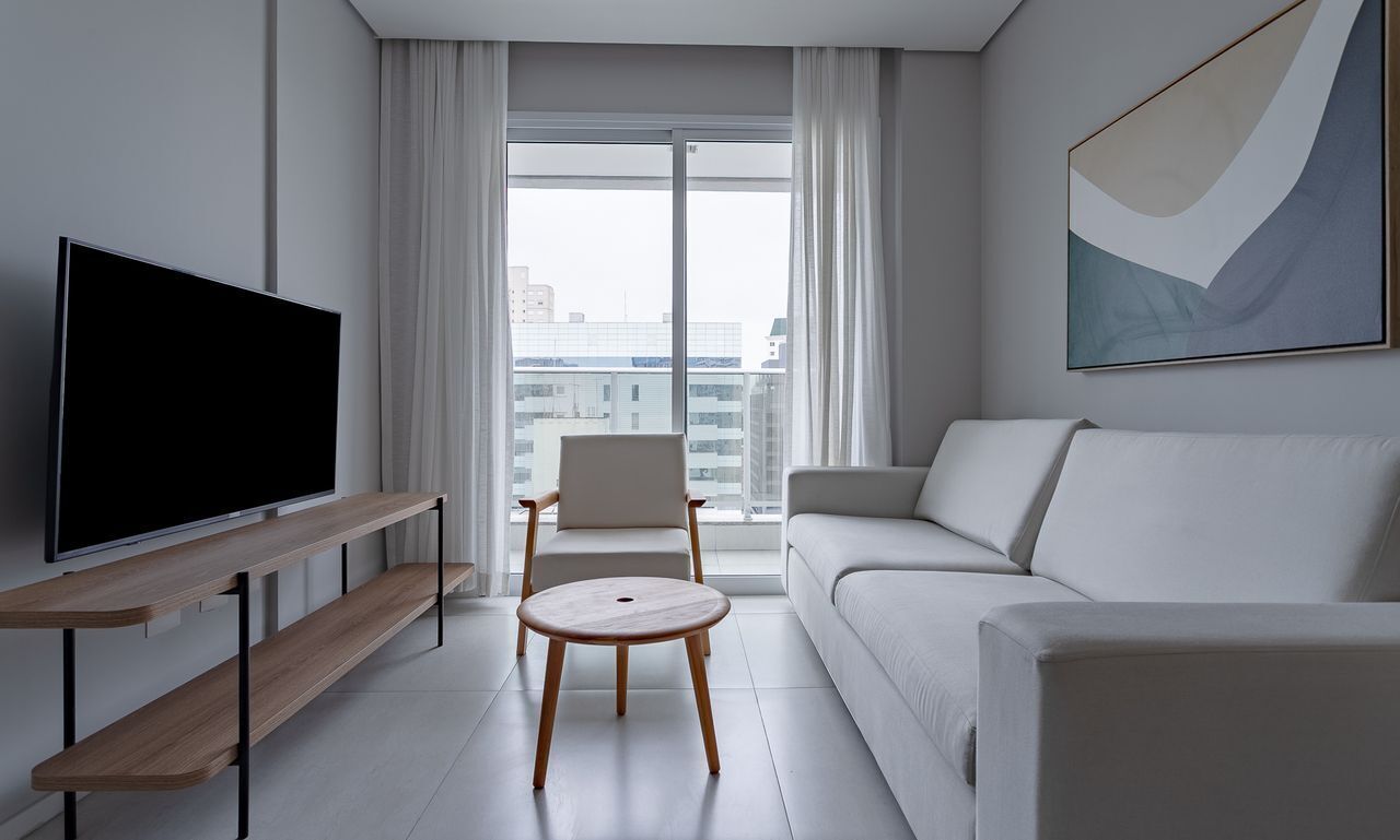 Un salon moderne avec un téléviseur à écran plat, un canapé, un fauteuil, une petite table ronde et une grande fenêtre avec des rideaux.