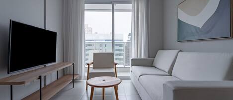 Un salon moderne avec un téléviseur à écran plat, un canapé, un fauteuil, une petite table ronde et une grande fenêtre avec des rideaux.