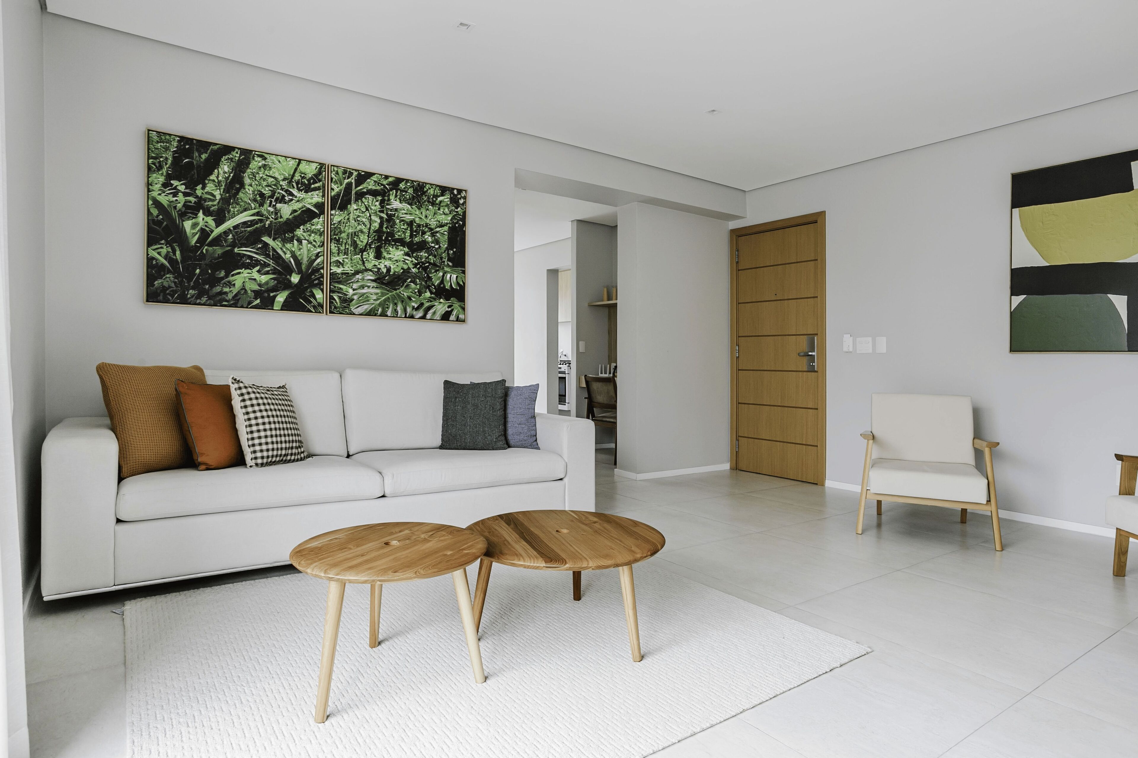 Una sala de estar moderna con un sofá blanco, dos mesitas de madera, un cuadro abstracto grande y una puerta de madera.