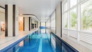 Uma piscina interior extensa, com azulejos azuis translúcidos, ladeada por janelas com persianas e vista para palmeiras lá fora.
