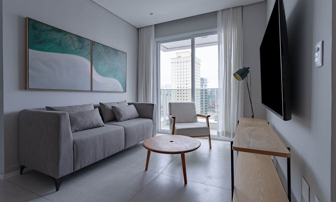 Uma sala de estar moderna com sofá, mesa de centro redonda, uma cadeira, televisão e uma grande janela com vista para a cidade.