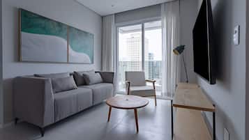 Uma sala de estar moderna com sofá, mesa de centro redonda, uma cadeira, televisão e uma grande janela com vista para a cidade.