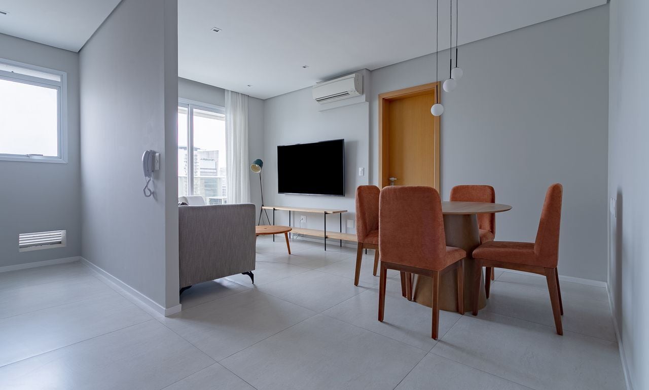 A modern living room with a dining area, a flat-screen TV mounted on the wall, and a large window providing natural light.