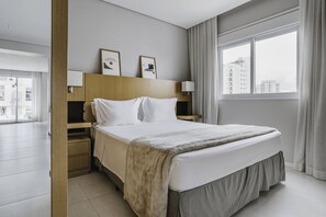 A modern hotel room with a large bed, a wooden headboard, two bedside tables with lamps, and a view of buildings through the window. - Tabas - GO 850 Building in Vila Olímpia (São Paulo)
