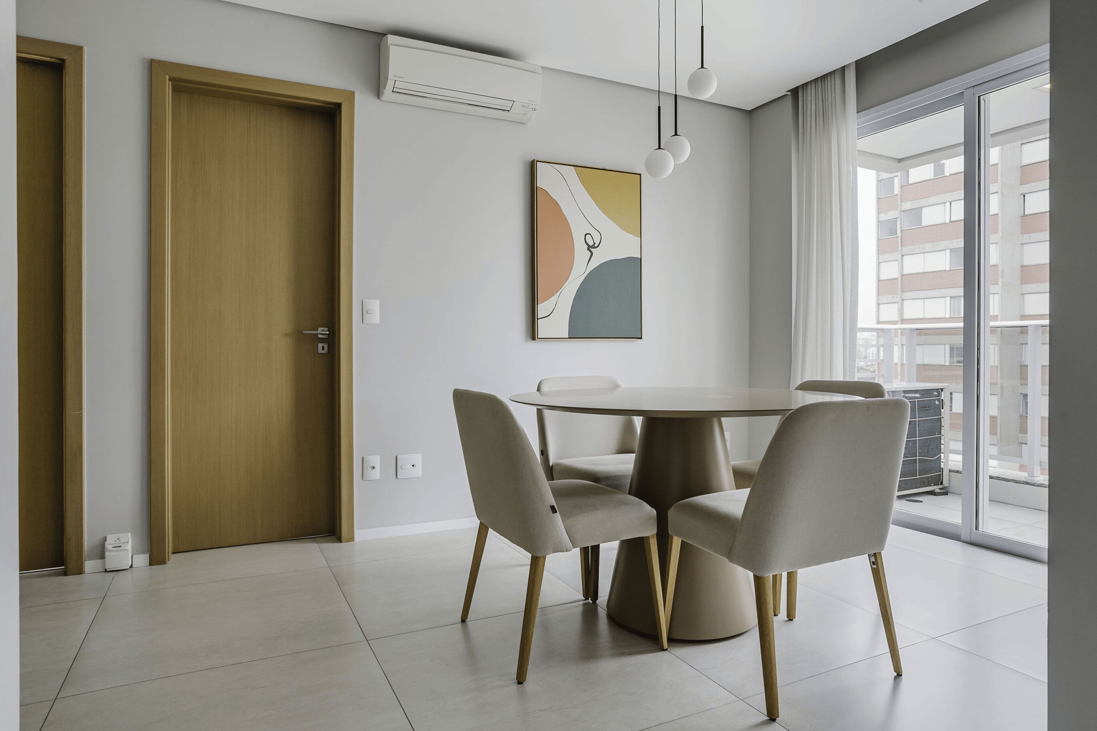 A modern dining area with a round table and four chairs, a wall-mounted air conditioner, and a large window with a view of buildings.