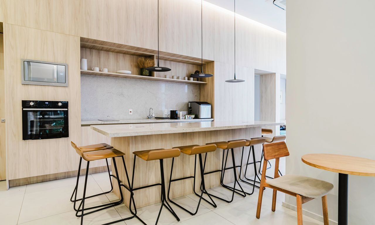 Cozinha moderna com forno embutido, micro-ondas e máquina de café. Há banquetas e uma mesa de madeira.