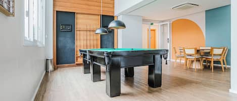 A modern indoor pool table with a wooden floor and pendant lighting, a wooden wall with a chalkboard, and a dining area with wicker chairs.