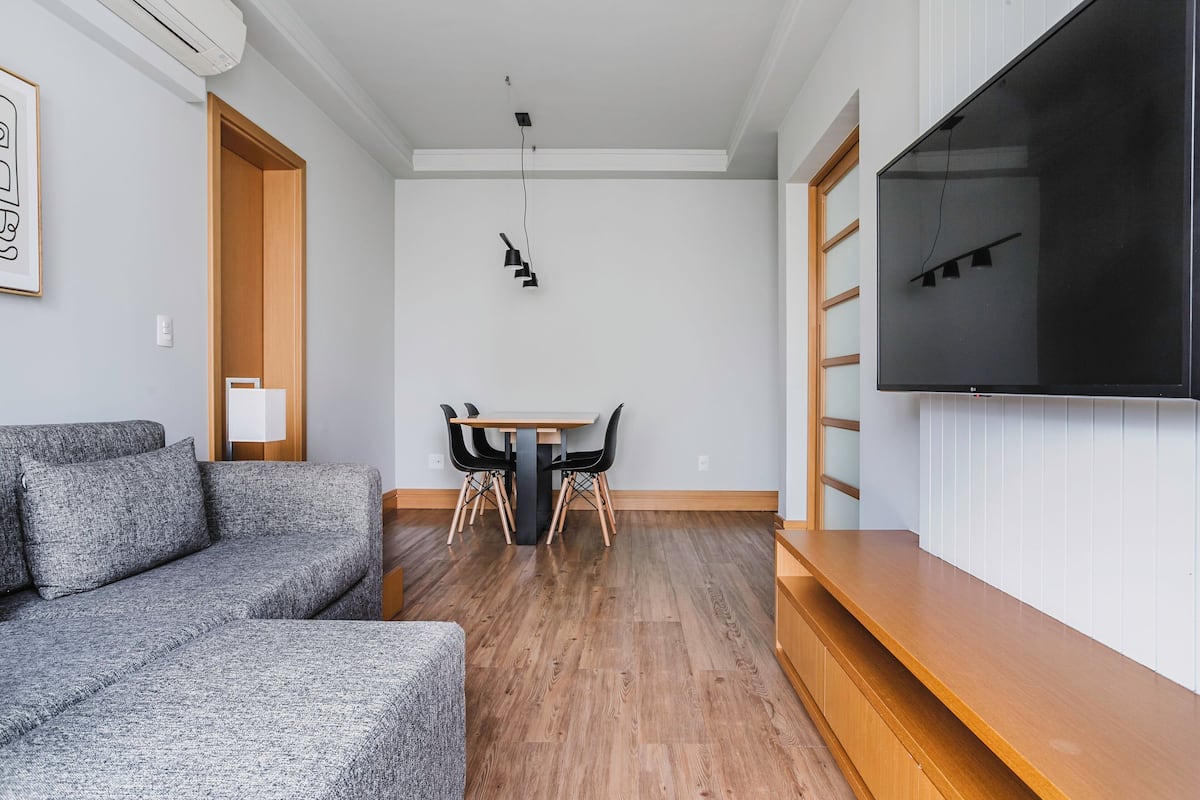 A modern living room with a grey sectional sofa, a wooden dining table with chairs, a flat-screen TV mounted on the wall, and a wooden bench.