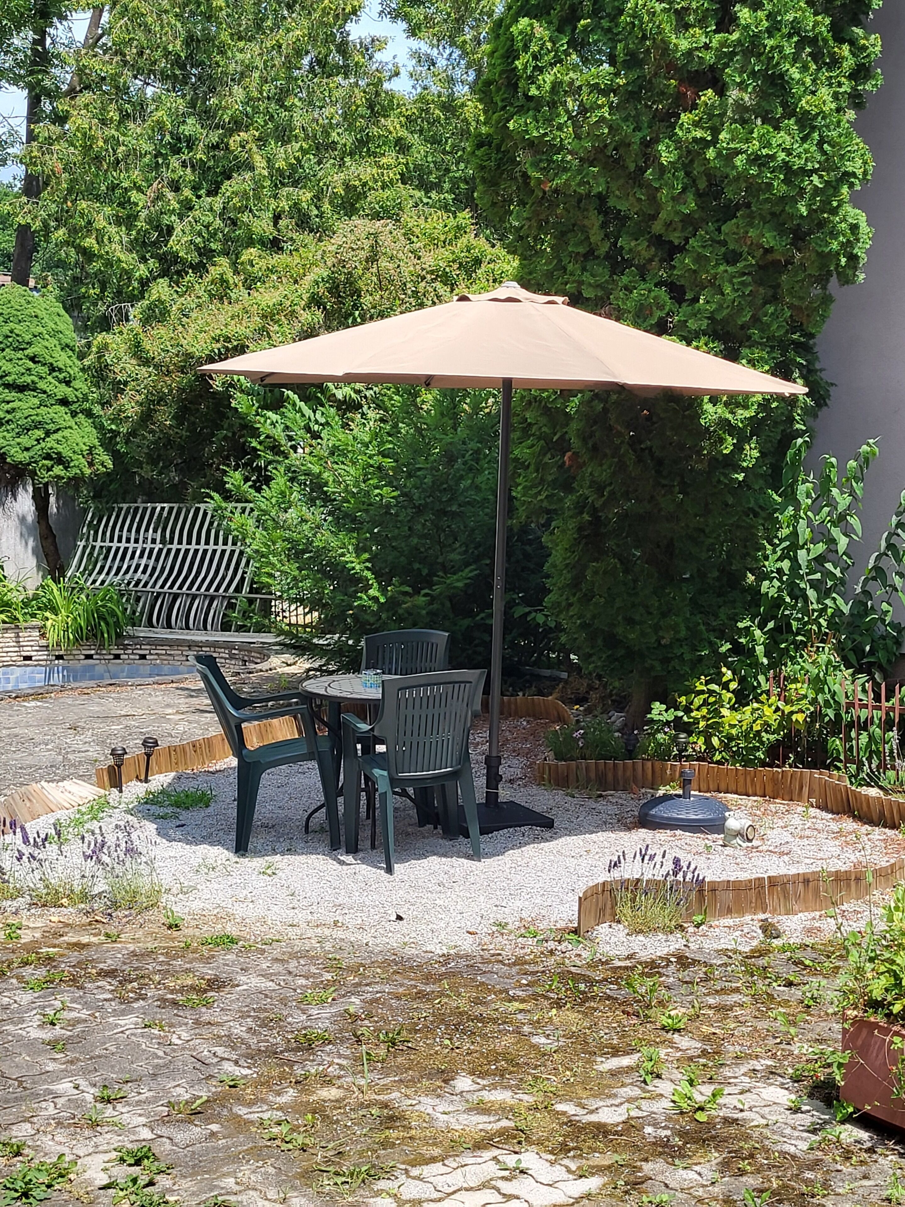 Een terras buiten met een tafel, stoelen, een grote parasol, een tuin met diverse planten en een houten omheining.