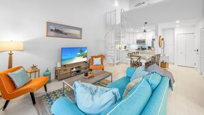 A spacious living area with a sofa, armchairs, a coffee table, and a TV. A kitchen is visible in the background with a dining area.