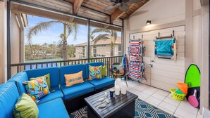 A sunlit balcony with a blue sofa, a ceiling fan, and a view of palm trees and houses.