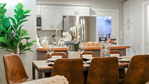 A dining area with a table set for four, surrounded by brown chairs. There is a potted plant to the left and a vase with white flowers on the table. The room has white cabinets and a kitchen area in the background. - Private hot tub / Close to town / Cozy and pet friendly (Chelan)