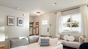 A modern living room with a large sectional sofa, a smaller sofa, and a bed with striped pillows. There are two framed artworks on the wall, a door, and a window with blinds. - Private hot tub / Close to town / Cozy and pet friendly (Chelan)