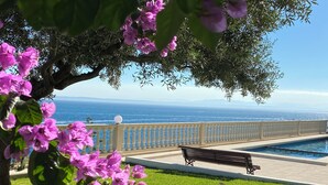 Un área junto a la piscina con un banco, vista al océano y un árbol de flores rosadas.