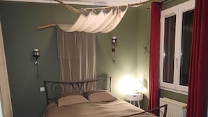 Une chambre à coucher avec un lit, une table de chevet, une lampe fixée au mur, une fenêtre avec des rideaux rouges et un radiateur.