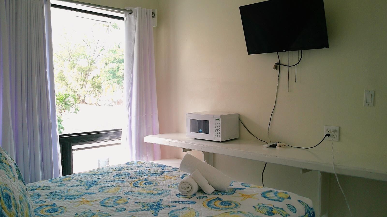 Habitación de hotel con cama, un microondas en un estante, televisor montado en la pared y una ventana con cortinas.