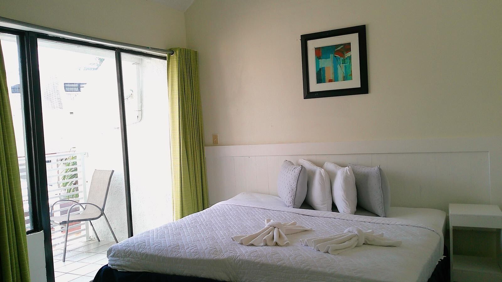 A hotel room with a bed, pillows, a framed picture on the wall, and a balcony with a chair.