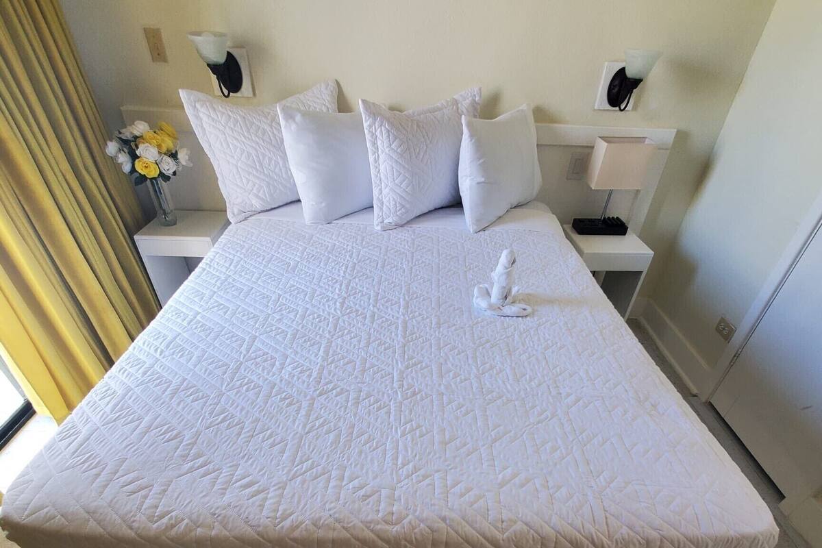 Una cama grande con ropa de cama blanca y un patrón acolchado, flanqueada por mesitas de noche blancas con lámparas, en una habitación con piso de baldosas y una cortina amarilla.