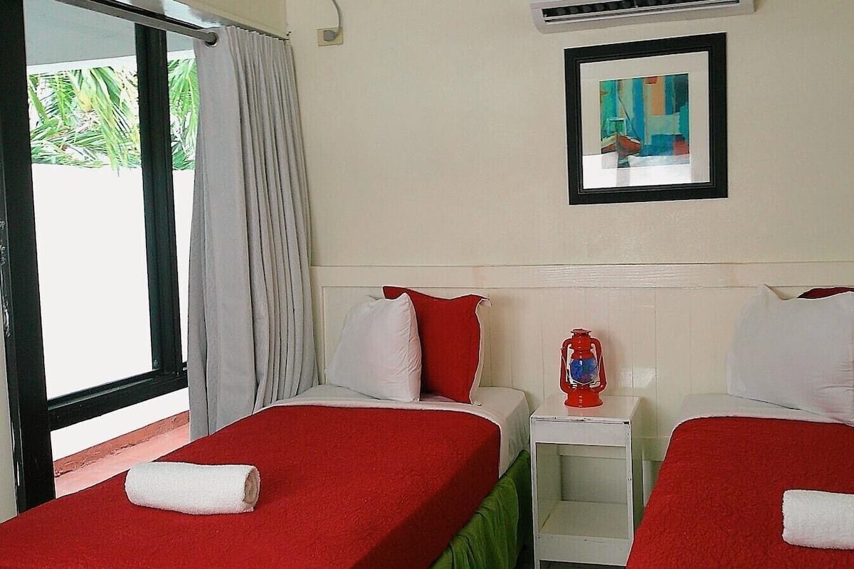Habitación de hotel con dos camas, cada una con ropa de cama roja y almohadas blancas, una mesita de noche pequeña con un farol rojo y un cuadro en la pared.