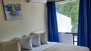 A hotel room with a bed, pillows, a painting, an air conditioner, and a window with curtains. - The Island Garden Hotel (Nassau)
