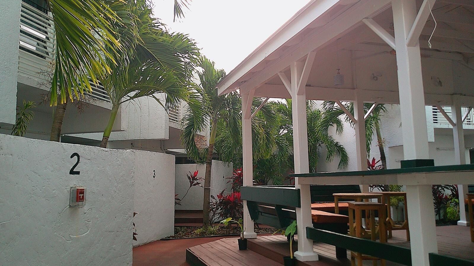 A covered outdoor seating area with wooden tables and benches, surrounded by white walls with numbers indicating room or unit designations.